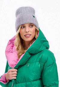 Zielona kurtka puchowa z różowym wnętrzem i dużym kołnierzem, o błyszczącym wykończeniu i zapięciu na zatrzaski. Na głowie szara dziergana czapka beanie.