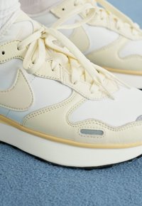 Nike copalke v beli in svetlo bež barvni shemi, iz usnja in tekstilnih materialov, s ploskimi vezalkami in oblazinjeno podplatom.