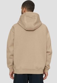 Felpa oversize beige con cappuccio spazioso e polsini a costine. Tessuto morbido, senza marchi visibili e vestibilità rilassata, vista da dietro.