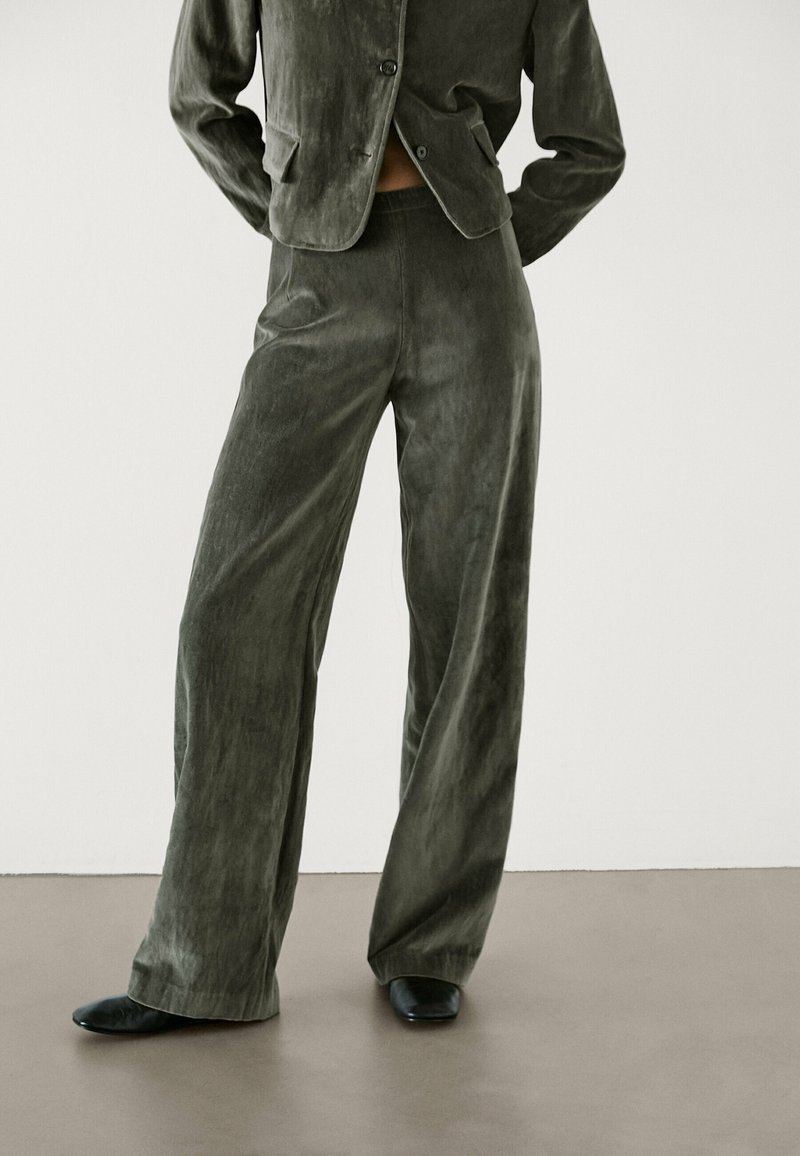 Personne portant un pantalon large couleur olive foncé et un blazer assorti, debout les mains dans le dos, chaussures noires visibles sur un sol neutre.