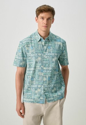 Bărbat purtând o cămașă cu nasturi cu mâneci scurte, având un model geometric tropical verde și albastru, combinată cu pantaloni de culoare bej deschis.