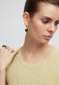 Orecchini pendenti color oro con una perla nera e un accento ovale di colore chiaro. Il modello indossa un maglione beige a trama.
