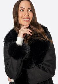 Abrigo negro con un lujoso cuello y puños de piel de peluche negra. Presenta una textura suave y un diseño estructurado. Detalles de canalé blanco en la muñeca.