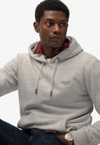 Lichtgrijze hoodie met trekkoorden, kangoeroezak en logo. Draagt over een rood geruit shirt, gecombineerd met donkere spijkerbroek en een gouden horloge.
