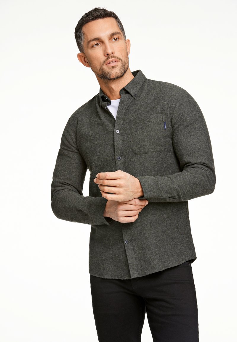 Donkergroene button-up shirt met een zak, gemaakt van textuurstof. Heeft lange mouwen en een afgeronde zoom. Draagt over een wit T-shirt.