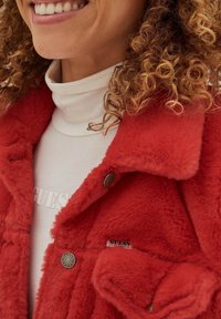 Rode fake bontjas met een kraag, drukknoopsluiting en zijzakken. Gedragen over een witte coltrui met een reliëftekstdetail.