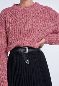 Rosa gestrickter Pullover mit strukturiertem Muster, Rundhalsausschnitt und überschnittenen Schultern, kombiniert mit einem schwarzen Faltenrock und einem schwarzen Gürtel.
