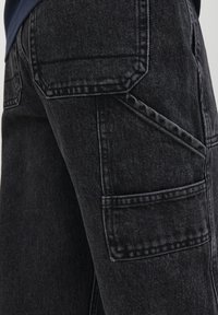 Svarta denim cargo-byxor med flera fickor, kontrasterande sömmar och en texturerad yta. Sidovy som visar fickdetaljer.