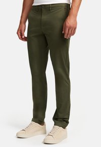 Chinos verde oscuro de tela suave, con cierre de botón y cremallera, bolsillos laterales y diseño de pierna estrecha, combinados con zapatillas beige.