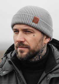 Gray knitted beanie with a ribbed texture and a brown leather patch. The wearer has a dark jacket and an open collar showing a black turtleneck.