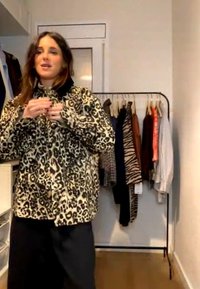 Γυναίκα με σακάκι με λεοπάρ μοτίβο στέκεται μπροστά σε μία κρεμάστρα ρούχων που κρατά διάφορα ρούχα με σχέδια και μονόχρωμα σε ένα δωμάτιο.
