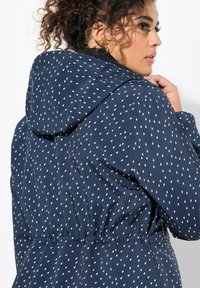 Giacca impermeabile blu navy con cappuccio, caratterizzata da un motivo a goccia bianca, vita elastica e tessuto liscio. Presenta un design moderno.