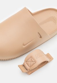 Bežiniai Nike šlepetės su lygiu plastiko viršumi ir tekstūruota vidine dalimi, kartu su atitinkamu bežiniu reguliavimo arba pritvirtinimo spaustuku.