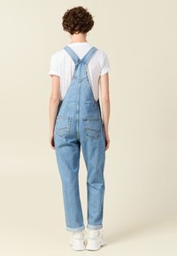 Un overol de mezclilla en azul claro con tirantes ajustables, bolsillos laterales y bolsillos traseros. Presenta un ajuste holgado y dobladillos con vuelta.