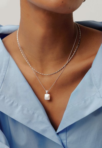 Δύο ασημένια κολιέ γύρω από τον λαιμό ενός ατόμου, το ένα με ένα μικρό τετράγωνο λευκό μαργαριτάρι ως μενταγιόν, φορώντας ένα ανοιχτό μπλε πουκάμισο με γιακά.