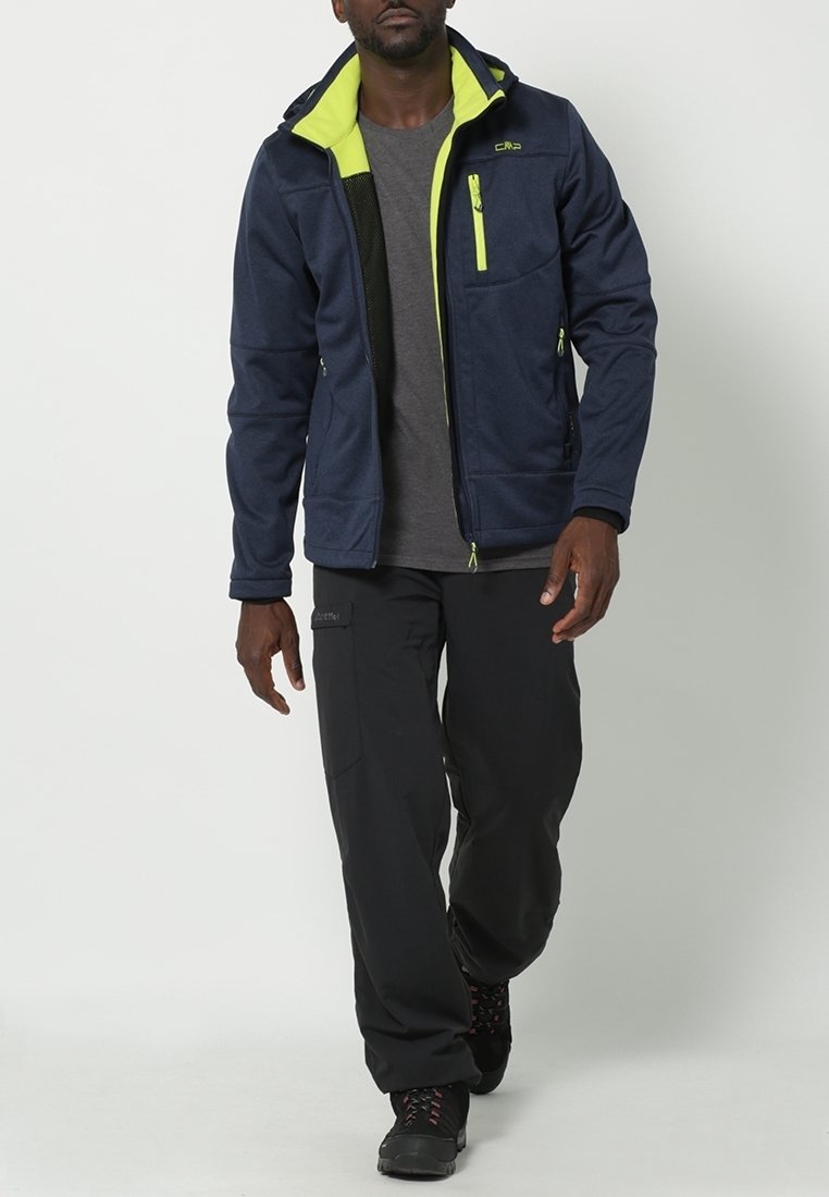 Homme marchant portant une veste bleu marine avec doublure jaune fluo, chemise grise, pantalon cargo noir et chaussures de randonnée noires.