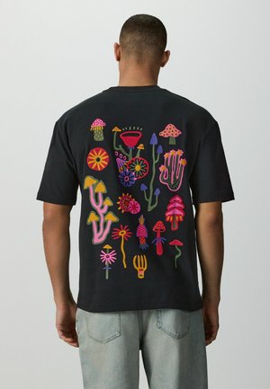 Mann trägt ein schwarzes T-Shirt mit bunten abstrakten Pilz- und Blumenillustrationen auf dem Rücken, kombiniert mit hellblauen Jeans.