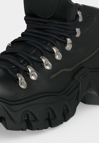 Μαύρο ανθεκτικό μποτάκι πεζοπορίας με χοντρή σόλα, μαύρα στρογγυλεμένα κορδόνια ασφαλισμένα με ασημένια μεταλλικά κρικάκια και ανάγλυφο λογότυπο στο πλάι.