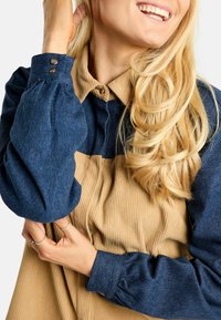 Farmerral és kordbársony felső galléros nyakkal, ballon ujjakkal és gombos részletekkel. Kék és barna színblokkolt kialakítás jellemzi.