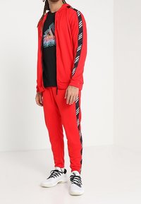 Homme portant une veste de survêtement rouge et un pantalon avec des détails en noir et blanc, un t-shirt graphique noir et des baskets blanches, se tenant dans un espace blanc.