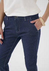 Jean en denim foncé avec un design ajusté, présentant des coutures contrastantes, des poches avant et une fermeture à bouton à la taille. Texture lisse.