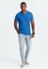 Pánské modré tričko henley s krátkým rukávem, džíny s ošoupanými detaily a světle hnědé tenisky. Jednoduchý, ležérní design.