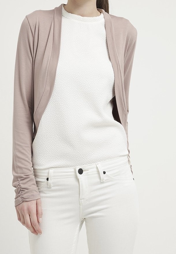 Jasno beżowy kardigan z miękkiego materiału dzianinowego, zapinany na otwarto, z długimi rękawami z marszczonym detalem, noszony na teksturowanej białej bluzce i białych dżinsach.