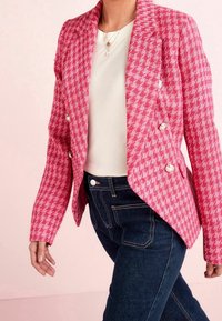 Růžový houndstooth blazer s přiléhavým střihem, dvouřadým zapínáním, texturovanou tkaninou a vyhrnutými klopami, kombinovaný s tmavými džíny.