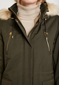 Femme portant une veste d'hiver vert olive avec des fermetures éclair dorées et une capuche bordée de fourrure, par-dessus un pull à col roulé beige, gros plan sur le torse.