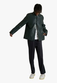 Giacca in eco-pelle verde lucido con colletto a punta, chiusura con bottoni a pressione e vestibilità ampia, abbinata a pantaloni scuri e sneaker bianche.