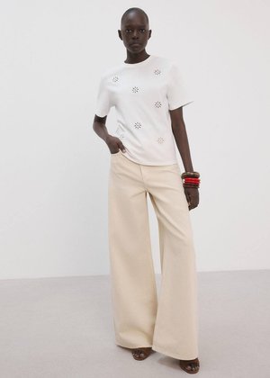 Persoană cu pielea închisă la culoare, purtând o cămașă albă cu modele florale decupate, pantaloni bej largi, brățări roșii și maro, stând în fața unui zid alb.