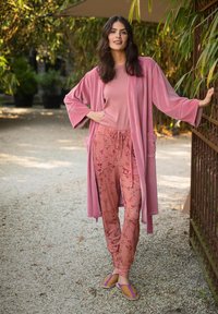 Woman standing outdoors wearing a pink striped top, pink floral pants, a pink robe, and tan sandals, leaning on a metal gate.