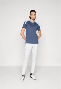 Navy short-sleeve sports shirt with light blue accents and red logo, paired with white pants and light-colored athletic shoes.