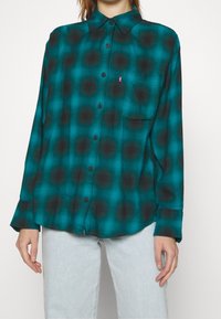 Camicia a maniche lunghe a quadri teal e nera con colletto abbottonato, taschino frontale e bottoni neri. Tessuto morbido con vestibilità comoda.