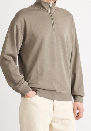 Beiger Halb-Zip-Sweatshirt mit gerippten Bündchen und Saum, mit kleinem mittig platziertem "HUGO"-Logo, getragen zu offwhite Hosen, Hand in der Tasche.