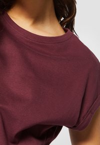 Bordové tričko s krátkým rukávem, kulatým výstřihem a měkkým bavlněným materiálem. Viditelné švy podél výstřihu a rukávů.