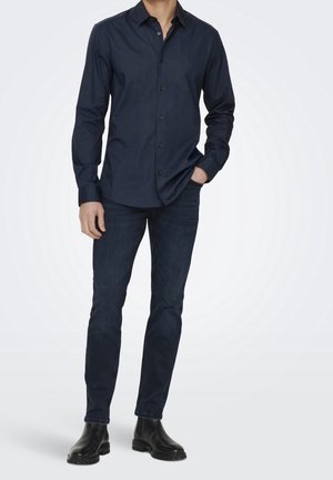 Camisa azul marino con botones, de corte entallado, mangas largas y textura suave, combinada con vaqueros azul oscuro y botas Chelsea negras.