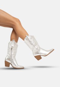 Botas de vaquero plateadas con un acabado brillante, caña texturizada, punta angulada y tacón de madera apilada. El cierre con cremallera lateral mejora la funcionalidad.