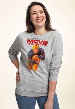 Lachende vrouw met een grijze trui waarop een vintage American footballspeler met bal staat afgebeeld en rode tekst 'MOVE THE CHAINS' aan de voorkant.