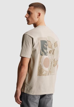 Mann mit kurzen Haaren trägt ein beiges T-Shirt mit abstraktem botanischem und geometrischem Aufdruck auf dem Rücken, kombiniert mit grauen Jeans.