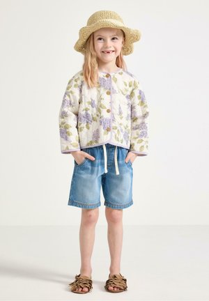 Ung pige smilende, iført en stråhat, blomsterprintet knapjakke, denimshorts og brune sandaler, står med hænderne i lommerne.