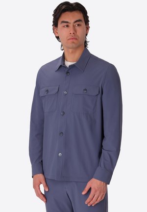 Uomo che indossa una camicia blu a maniche lunghe abbottonata con due tasche sul petto, in piedi e che guarda di lato su uno sfondo semplice.