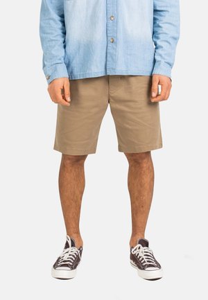 Hombre vestido con camisa celeste de botones, pantalones cortos beige hasta la rodilla y zapatillas negras de caña baja con cordones blancos, posando frente a un fondo blanco.