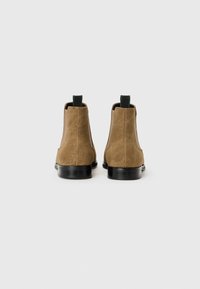 Botas Chelsea de ante marrón con una silueta elegante, que presentan una suela de goma negra y tiradores en verde oscuro en la parte posterior para ponérselas fácilmente.