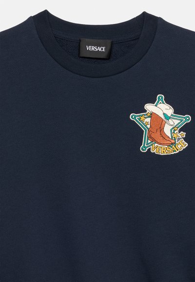 Μπλε σκούρο φούτερ Versace με πινελιά ενός καουμπόικου μπότες και καπέλου μέσα σε αστέρι, με κίτρινα αστέρια και τη λέξη "Versace" από κάτω.