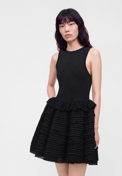 Femme aux longs cheveux noirs porte une robe noire sans manches avec un haut moulant et une jupe à volants superposés texturés, debout devant un fond uni.