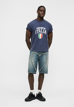 Μπλούζα από βαμβάκι σε ναυτικό μπλε με λευκό κείμενο και γραφικό της σημαίας της Ιταλίας, συνδυασμένη με ανοιχτό μπλε τζιν σορτς με φθορές σε σχήμα καρδιάς και μαύρα αθλητικά παπούτσια.
