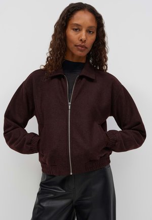 Femme aux cheveux bouclés portant une veste marron foncé zippée et un pantalon en cuir noir sur un fond clair uni.