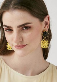 Boucles d'oreilles dorées avec des designs floraux complexes et une pierre noire centrale, suspendues à un support ondulé.