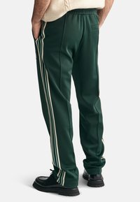 Zielone spodnie sportowe z białymi paskami po bokach, elastycznym paskiem w talii i tylną kieszenią. Materiał wydaje się gładki i lekko błyszczący. Widać czarne buty.